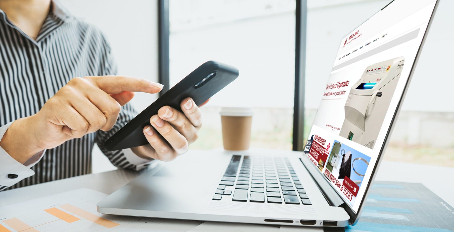 Business setting. A man's hands holding a operating a cellphone over a laptop with the IMEB website on the screen.